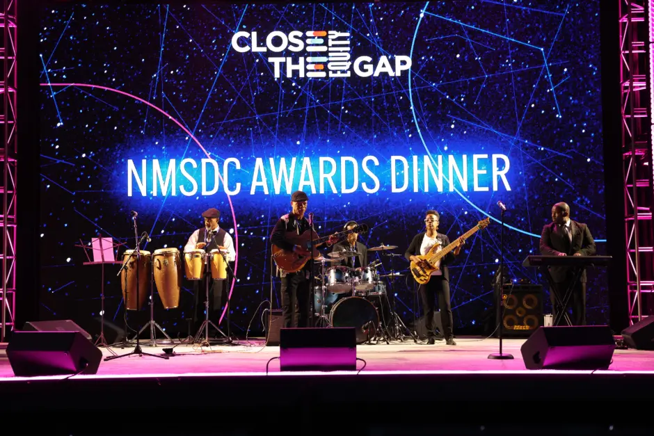 파란색과 검은색 화면 앞에서 밴드가 NMSDC 시상식 디너라고 적힌 음악을 연주합니다. 
