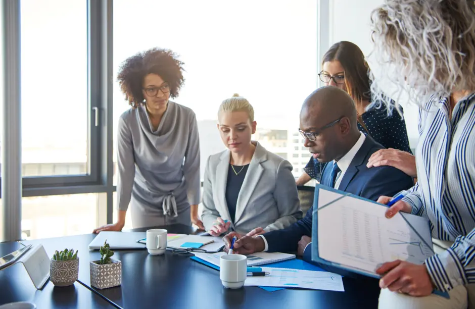 Image Description: Several individuals of diverse races standing over a table and working together in a business setting. 