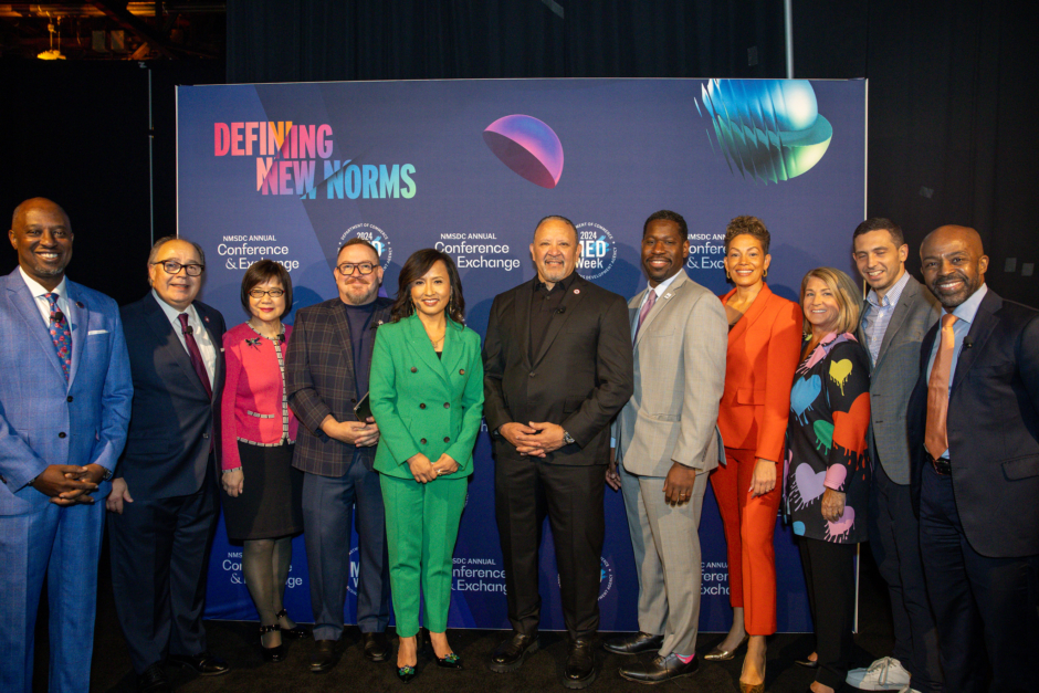 Diverse picture of individuals from all backgrounds with different colored suits against a colorful conference backdrop. 