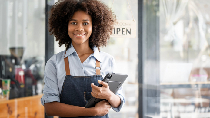 Retrato,de,un,joven,sonriente,afroamericano,barista,con,una,tableta,en,la,mano Mujer negra con delantal sosteniendo una tableta delante de una cafetería.