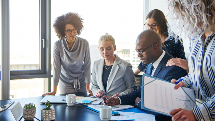 Group,Of,Focused,Businesspeople,Standing,Around,Their,Manager,Sitting,At Image Description: Several individuals of diverse races standing over a table and working together in a business setting.